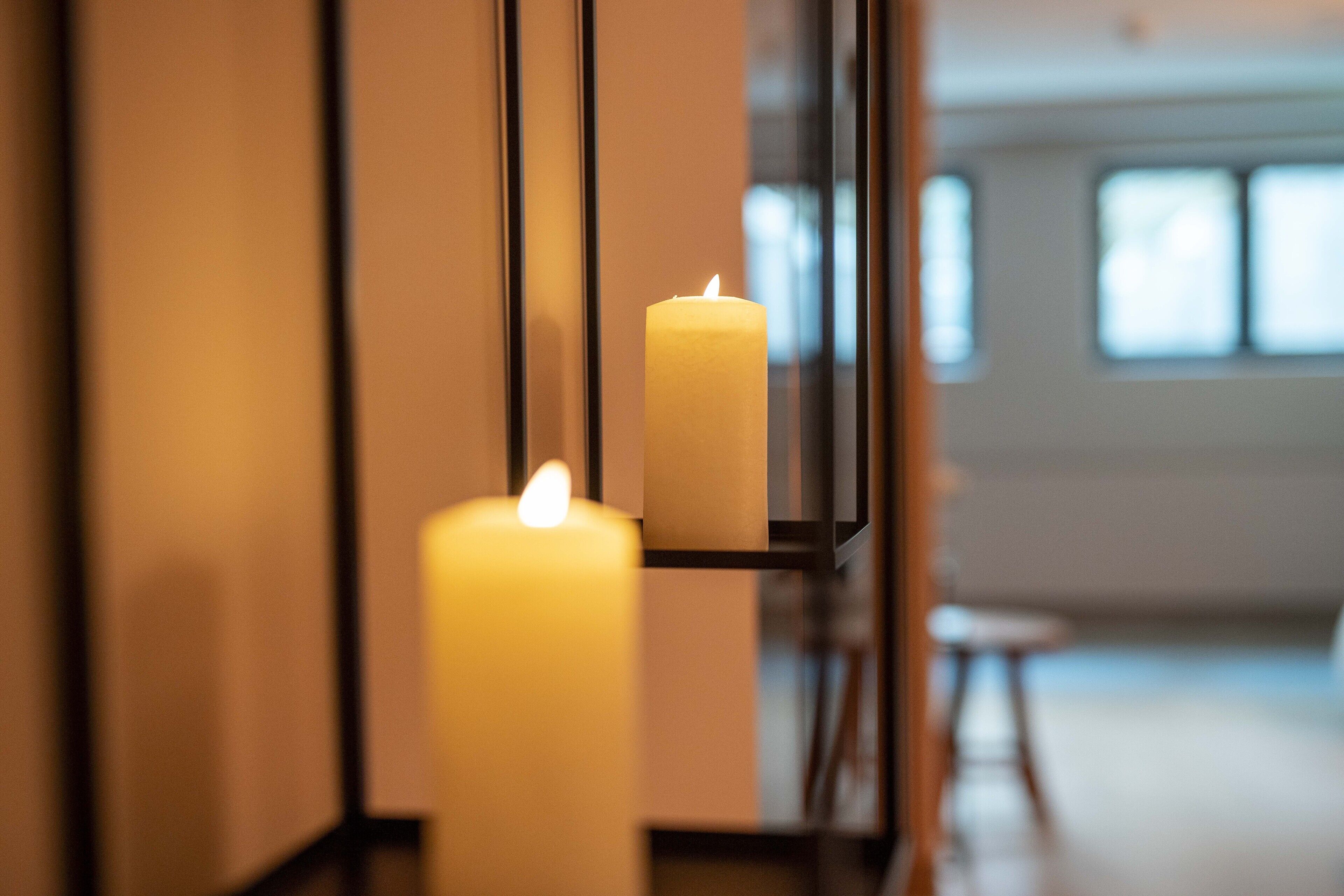 A candle on a shelf in a room with a window and a chair.