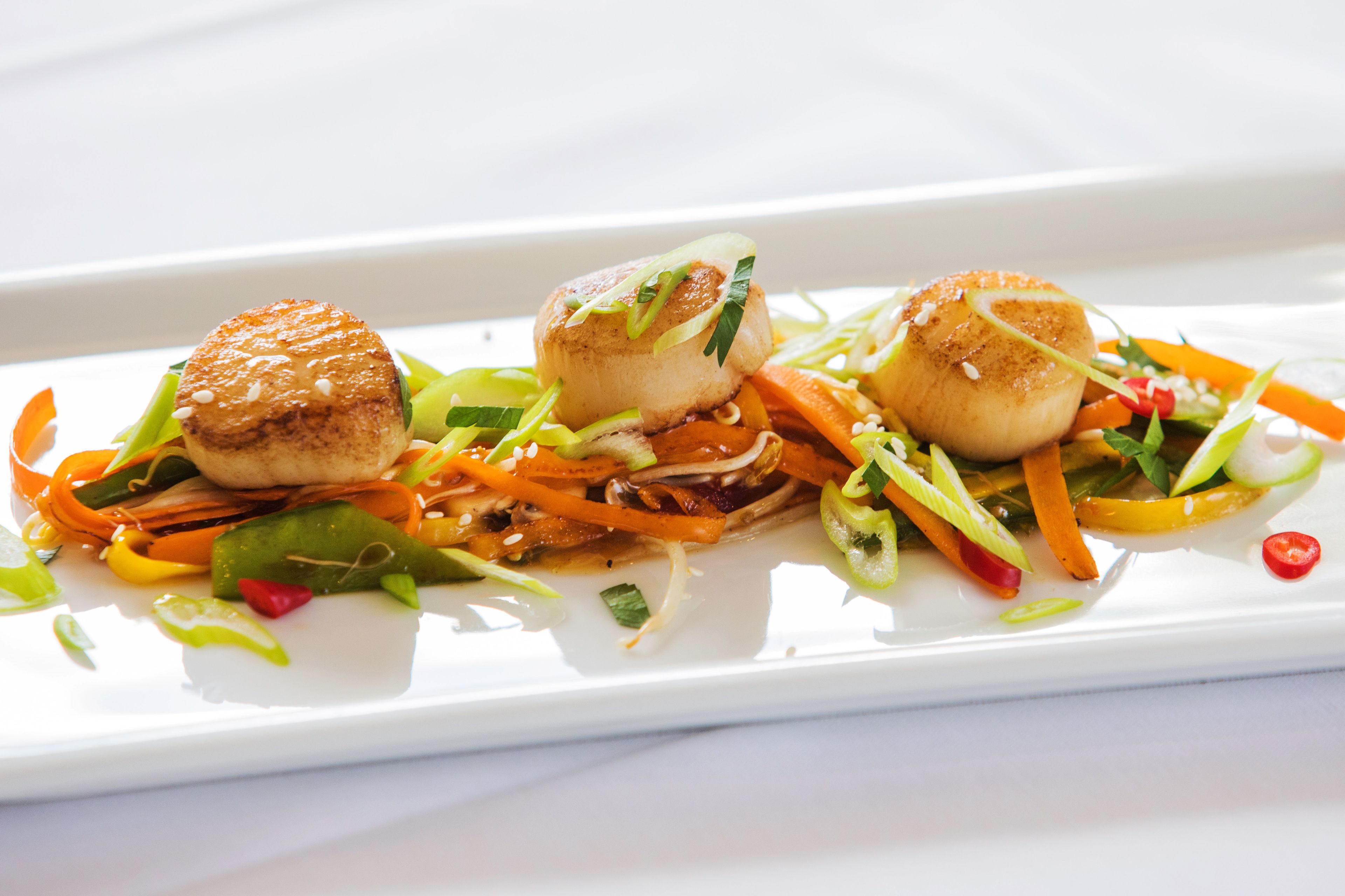 A white rectangular plate with three scallops and a variety of vegetables.
