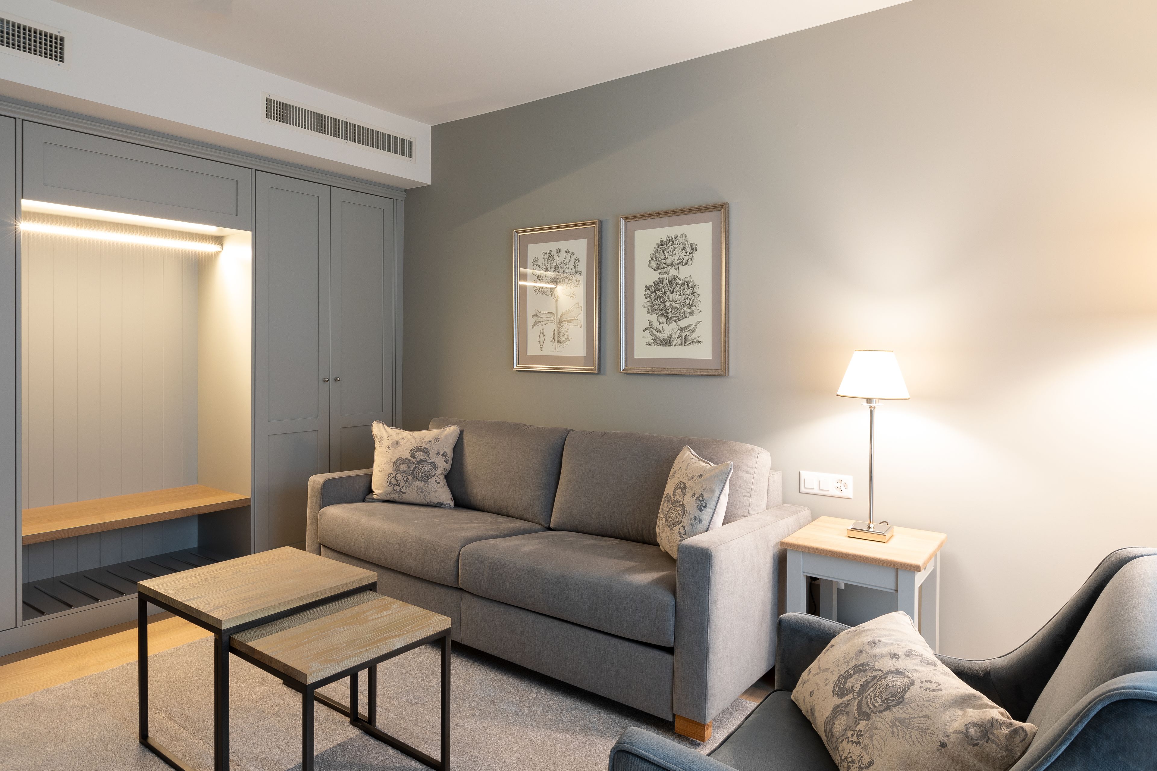 A modern living room with a grey sofa, a wooden coffee table, a side table with a lamp, and a bench. There are two framed botanical prints on the wall.