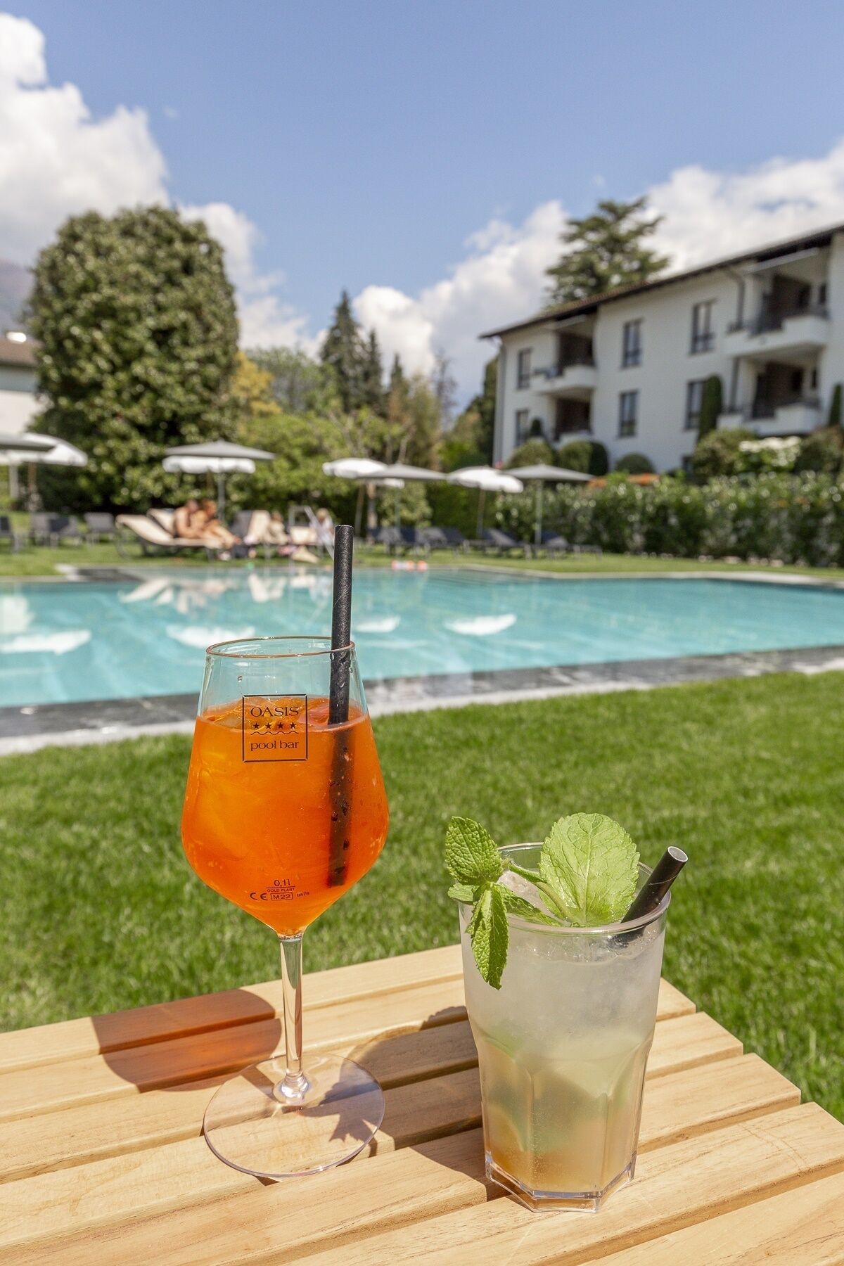 A glass of orange cocktail and a mint-infused drink on a wooden table by a poolside with a building in the background.