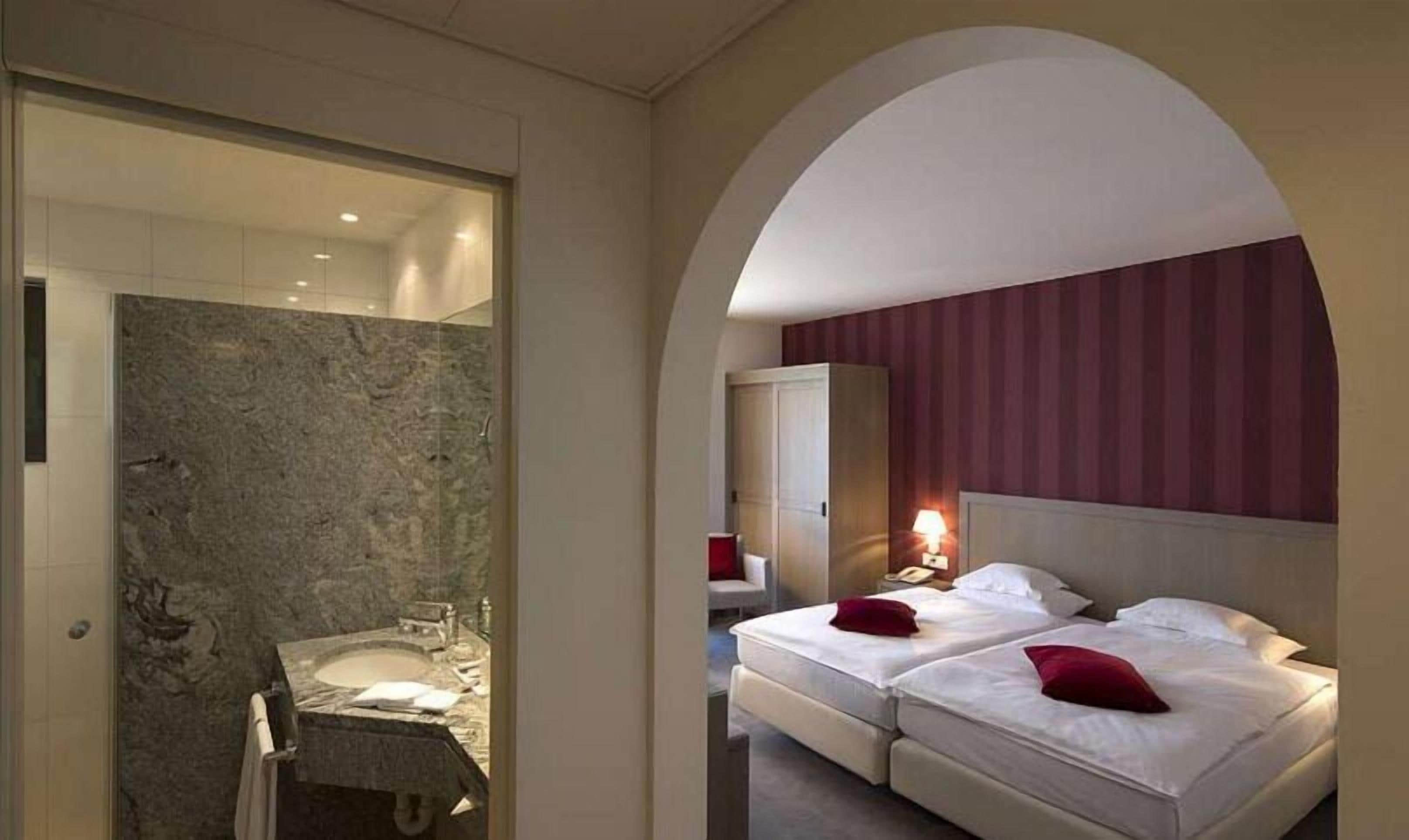 Habitación de hotel con baño que incluye un lavamanos y ducha de mármol, dos camas con almohadas rojas y un armario.