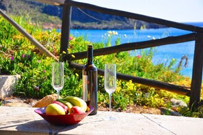 A table with a bowl of fruit, a bottle, and two glasses, set against a scenic coastal backdrop. - Coral Rooms (Sitia)