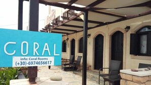 Uma área de estar coberta ao ar livre, com piso de pedra e mobiliário de madeira. Uma placa com o nome 'CORAL' e informações de contato é visível.
