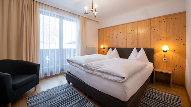 A modern hotel room with a large bed, two bedside lamps, a wooden headboard, a black armchair, and a view of the outdoors through sheer curtains. - Haus Margareth (Wagrain)