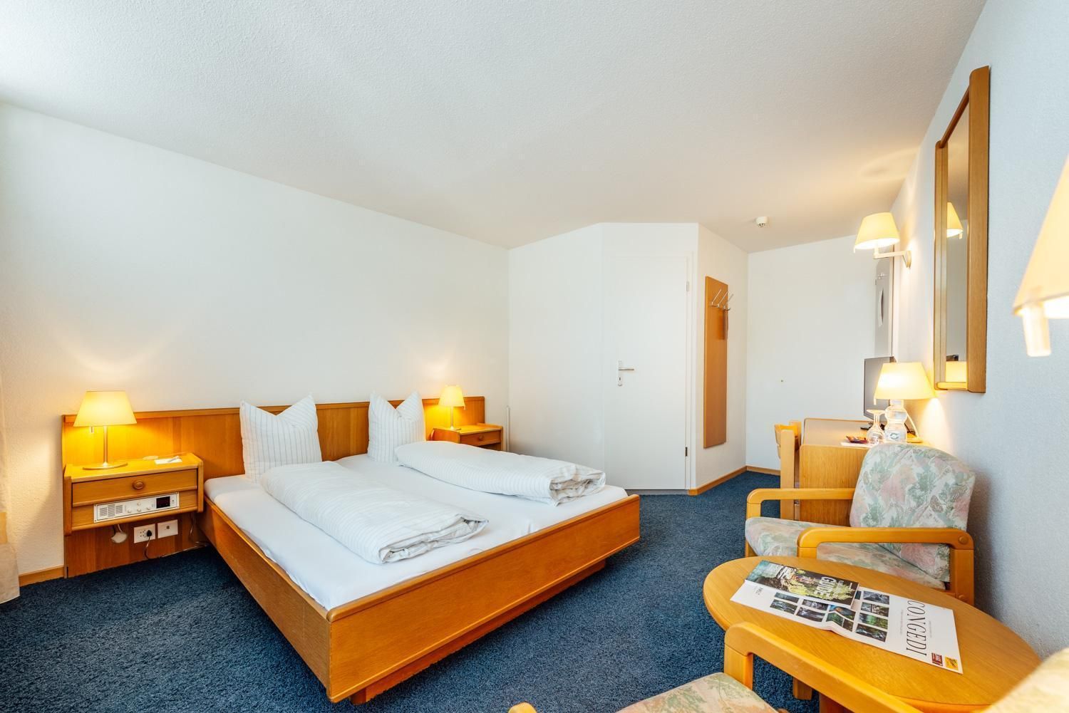 Ein Hotelzimmer mit einem Doppelbett, zwei Nachttischen mit Lampen, einem Sessel, einem kleinen Tisch mit Broschüren und einem Spiegel an der Wand.
