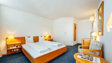Une chambre d’hôtel avec un lit double, deux tables de chevet avec des lampes, une chaise, une petite table avec des brochures et un miroir au mur.