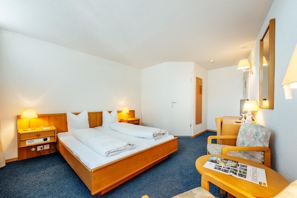 Une chambre d’hôtel avec un lit double, deux tables de chevet avec des lampes, une chaise, une petite table avec des brochures et un miroir au mur.