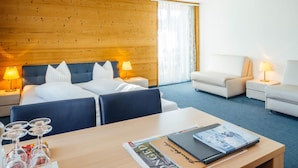 A hotel room with a wooden headboard, a bed with white linens, two bedside tables with lamps, a sofa, a small table with a magazine, and a window with curtains.