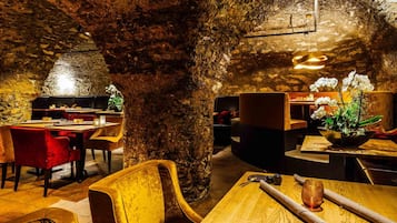 A restaurant with a rustic interior featuring exposed stone walls, wooden tables, and red upholstered chairs.