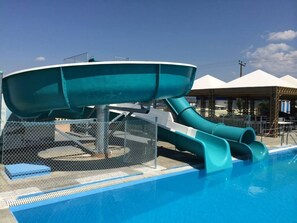 Une piscine avec deux toboggans, une zone délimitée par une clôture et un espace de repos couvert à l’arrière-plan.