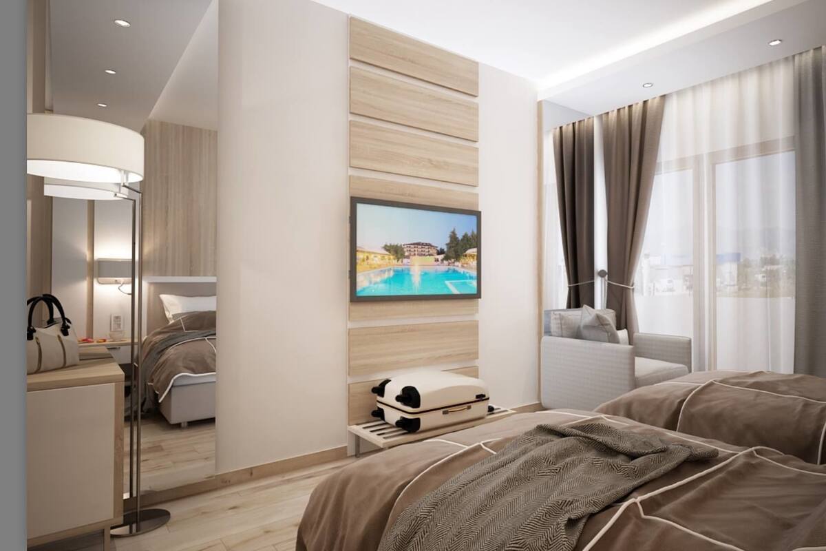 Quarto de hotel moderno com uma cama grande, uma escrivaninha com luminária, televisão na parede e vista para o exterior.