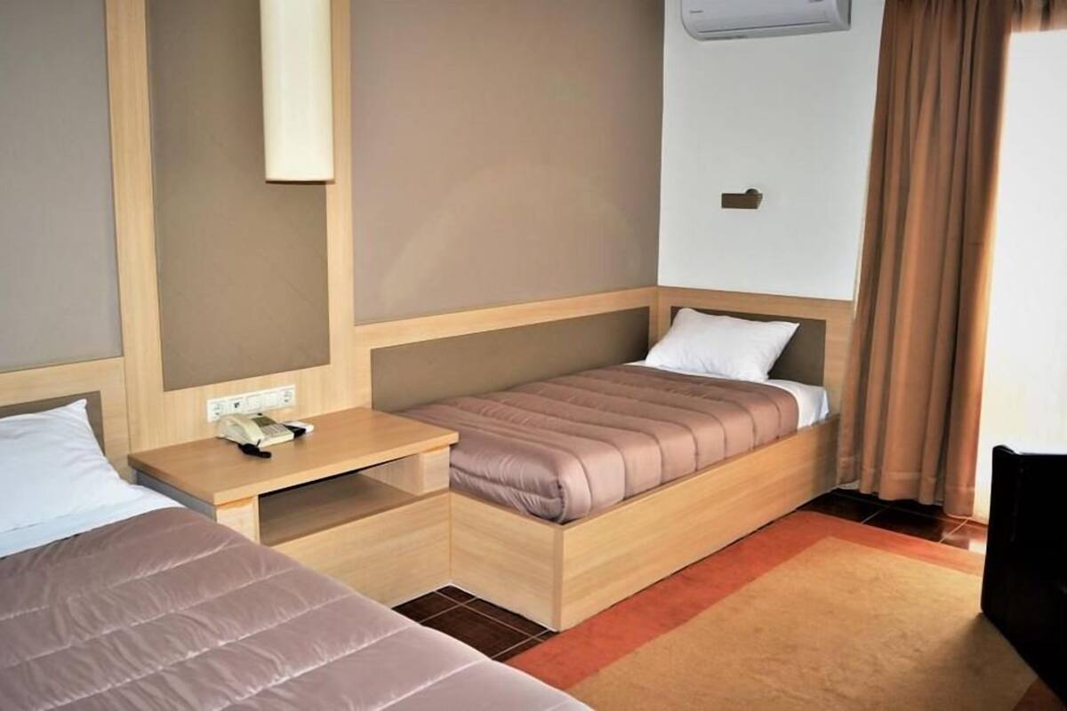 Quarto de hotel pequeno com duas camas, uma mesa de cabeceira de madeira, um telefone e uma janela com cortinas.