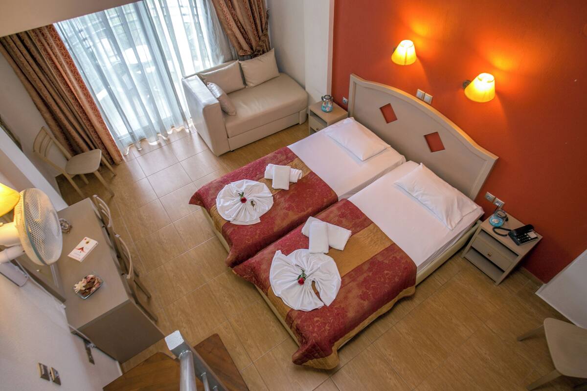Ein Hotelzimmer mit zwei Betten, einem Schlafsofa, einem Schreibtisch mit Ventilator und einem Fenster mit Vorhängen.