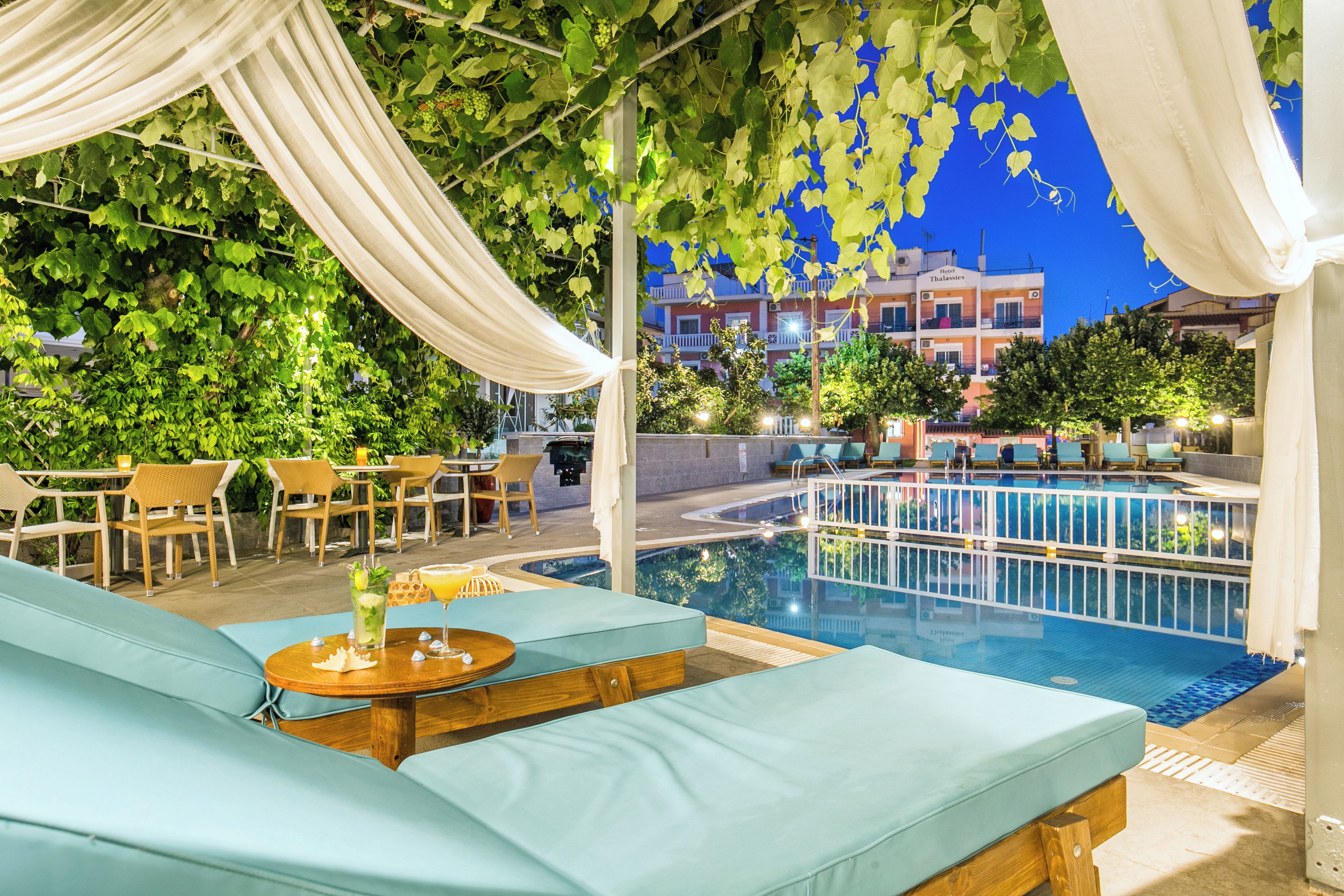 An outdoor pool area with lounge chairs and a table with drinks, surrounded by greenery and buildings in the background.