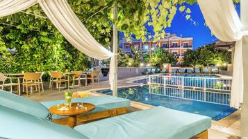 An outdoor pool area with lounge chairs and a table with drinks, surrounded by greenery and buildings in the background.