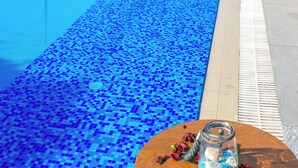 A swimming pool with blue mosaic tiles, surrounded by lounge chairs and a table with a glass jar. In the background, there are multi-story buildings with balconies and a hotel named "Hotel Thalassies." - Thalassies Nouveau (Thasos)