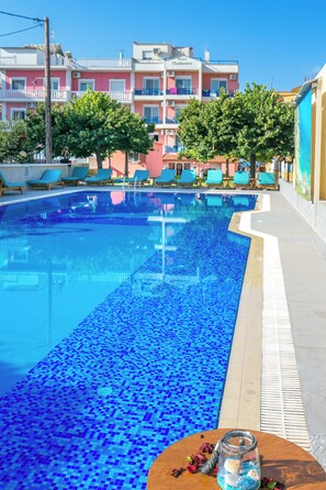 Ein Swimmingpool mit blauen Mosaikfliesen, umgeben von Liegestühlen und einem Tisch mit einem Glas. Im Hintergrund sind mehrstöckige Gebäude mit Balkonen und ein Hotel namens "Hotel Thalassies" zu sehen.