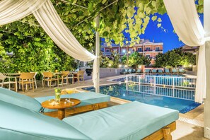 Area piscina esterna con lettini e un tavolo con bevande, immersa nel verde e con edifici sullo sfondo.
