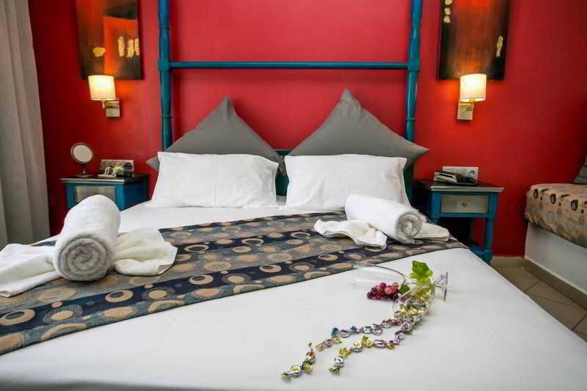 A hotel room with a red accent wall, a bed with white linens, two pillows, a patterned bedspread, and two bedside tables with lamps.