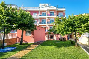 A hotel with a pink facade, multiple balconies, and a well-maintained garden with trees and a small fountain. - Thalassies (Thasos)
