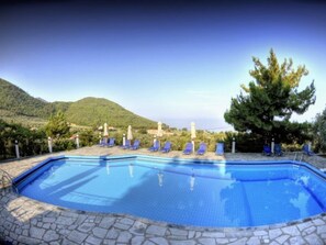 A swimming pool with stone tiles, surrounded by blue lounge chairs and umbrellas, set against a mountainous backdrop.