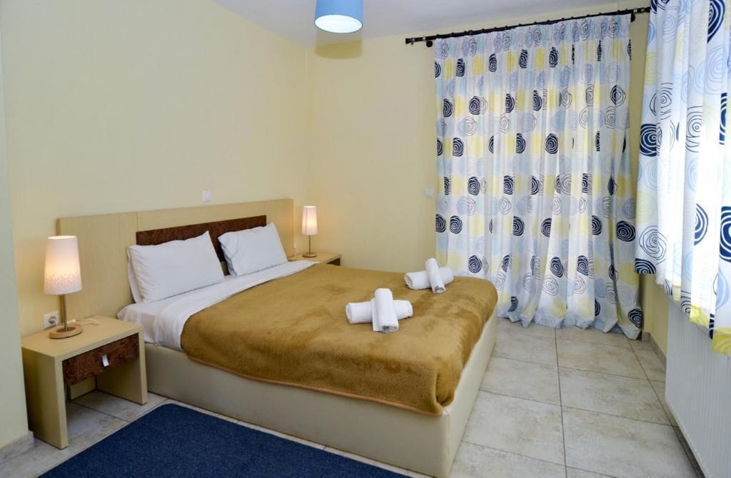 Habitación de hotel con cama, dos lámparas de noche, una mesita de noche, alfombra azul y cortinas estampadas.