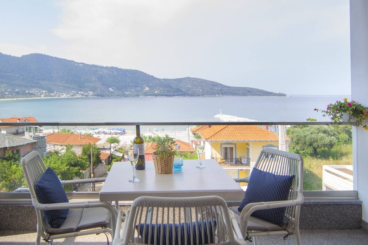 Un balcon doté de fauteuils et d’une table en osier, offrant une vue sur la côte, avec des bâtiments et des collines en contrebas.