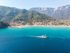 A coastal town with a boat on the turquoise sea, surrounded by mountains and houses.
