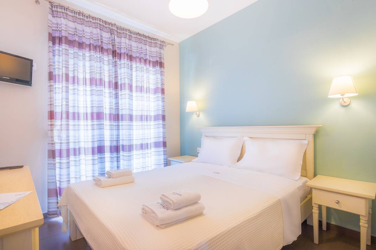 Un lit bien fait, avec du linge de lit blanc et deux serviettes enroulées, une table de chevet, une lampe fixée au mur et un téléviseur accroché au mur.