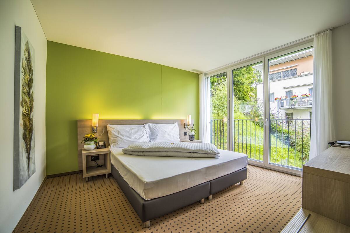 A bedroom with a bed, bedside table, lamp, and a view of a balcony.