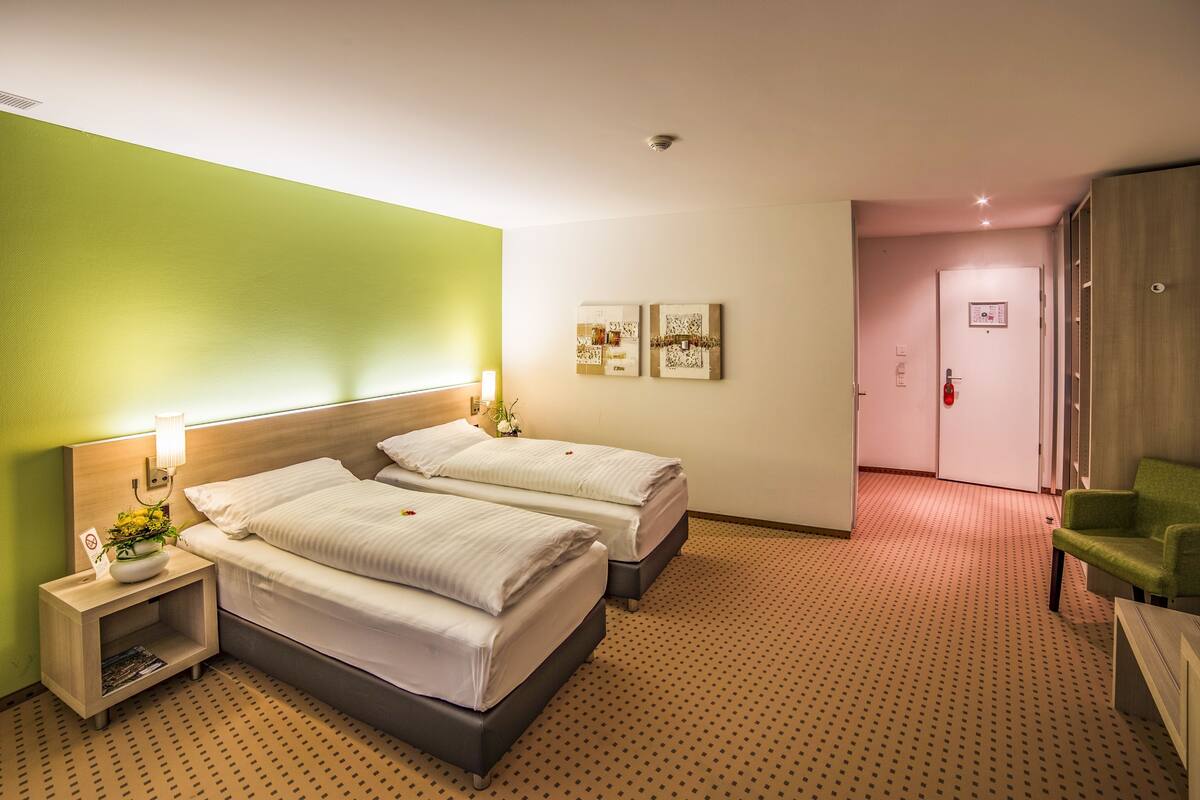 A hotel room with two beds, a green accent wall, a patterned carpet, and a green armchair.