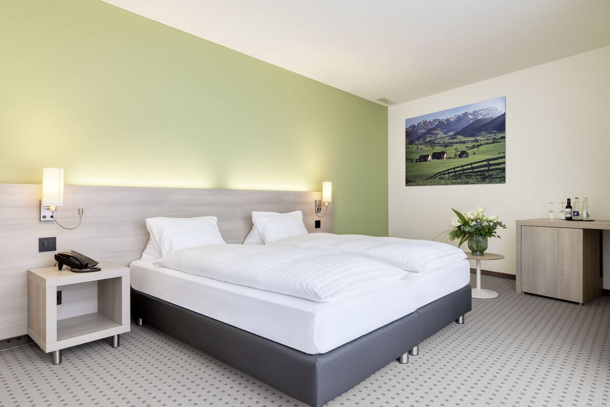 A modern hotel room with a large bed, bedside tables, a desk, and a wall-mounted painting of a mountain landscape.