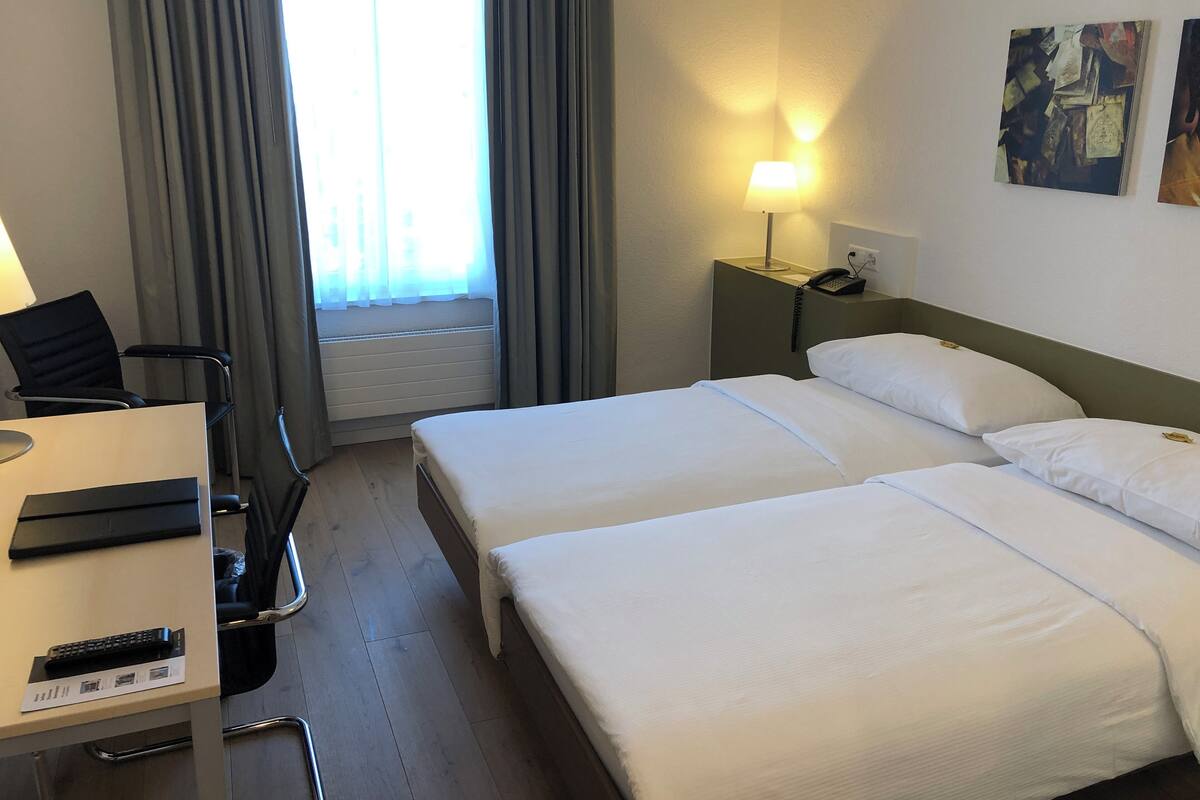A hotel room with two beds, a desk with a chair, a lamp, and a window with curtains.