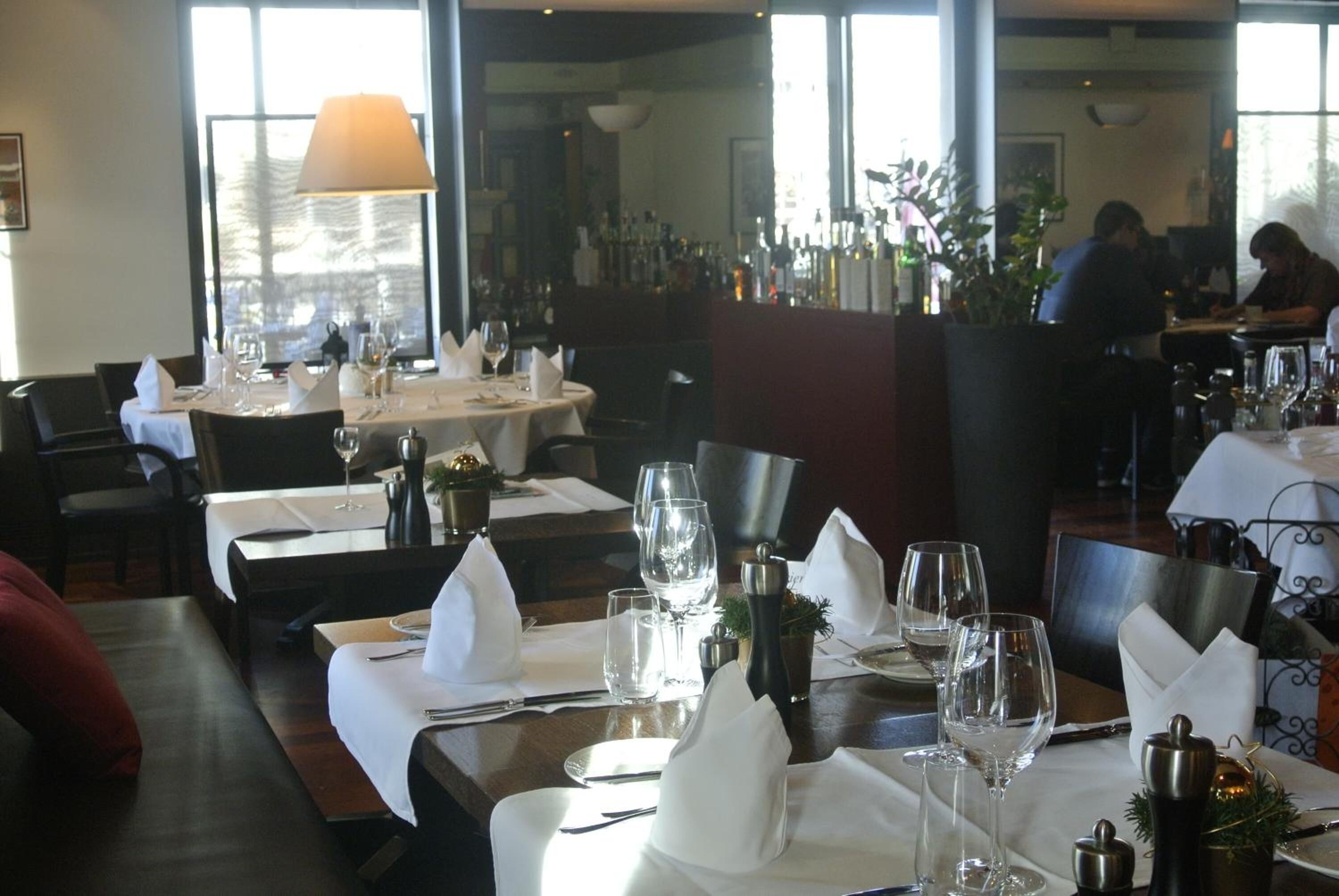 Een restaurant met gedekte tafels, witte tafellakens, wijnglazen en een groot raam op de achtergrond.