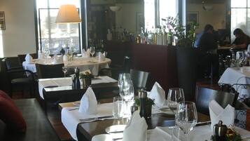 Een restaurant met gedekte tafels, witte tafellakens, wijnglazen en een groot raam op de achtergrond.