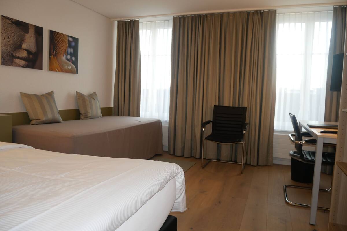 A hotel room with two beds, a desk, a chair, and a large window with curtains.