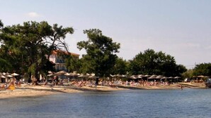 A beach with umbrellas and people, trees, and a building in the background. - Sunrise Beach (Thasos)