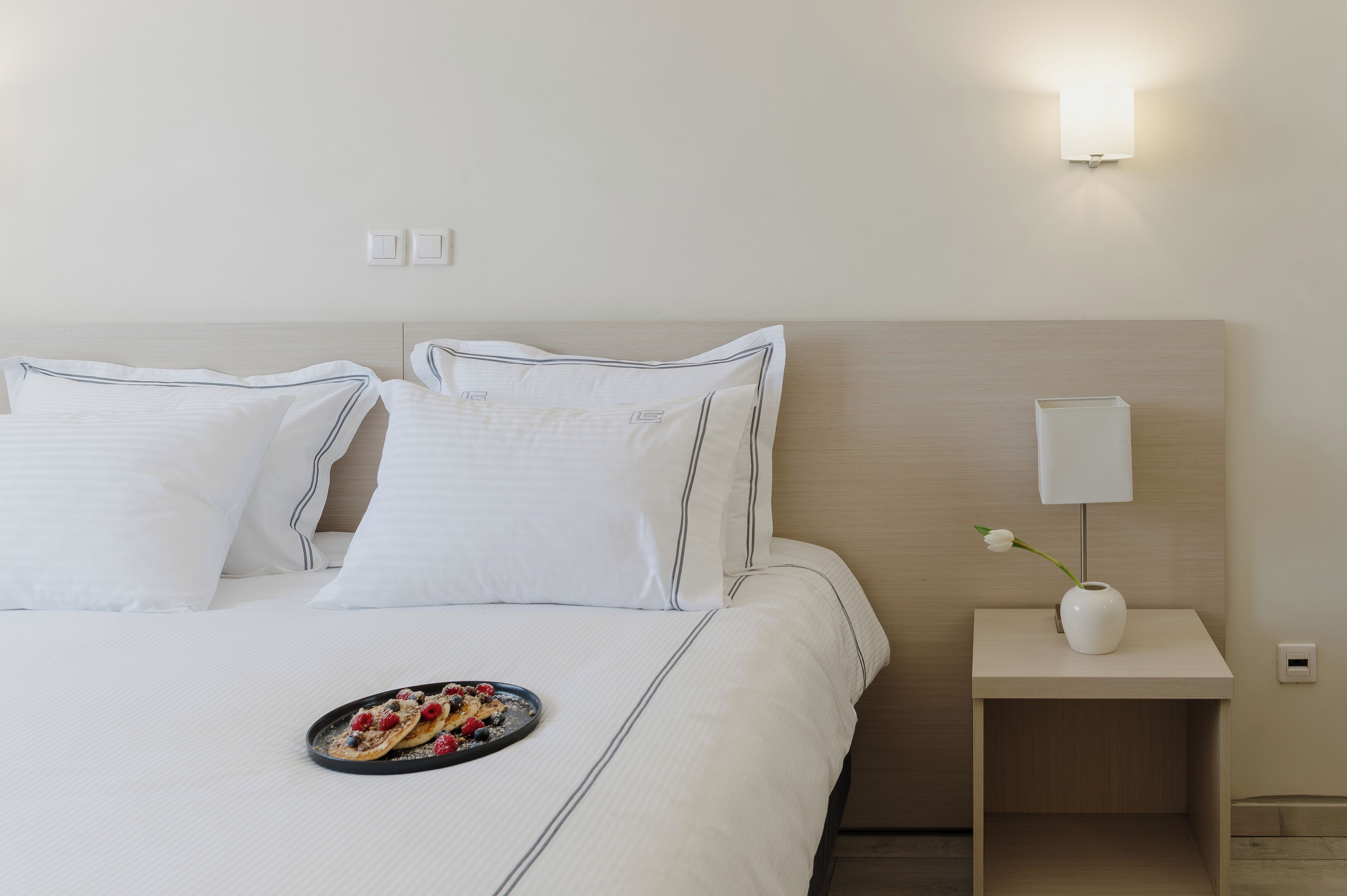 A neatly made bed with white linens and striped pillows, a bedside table with a vase and a lamp, and a plate of fruit on the bed.