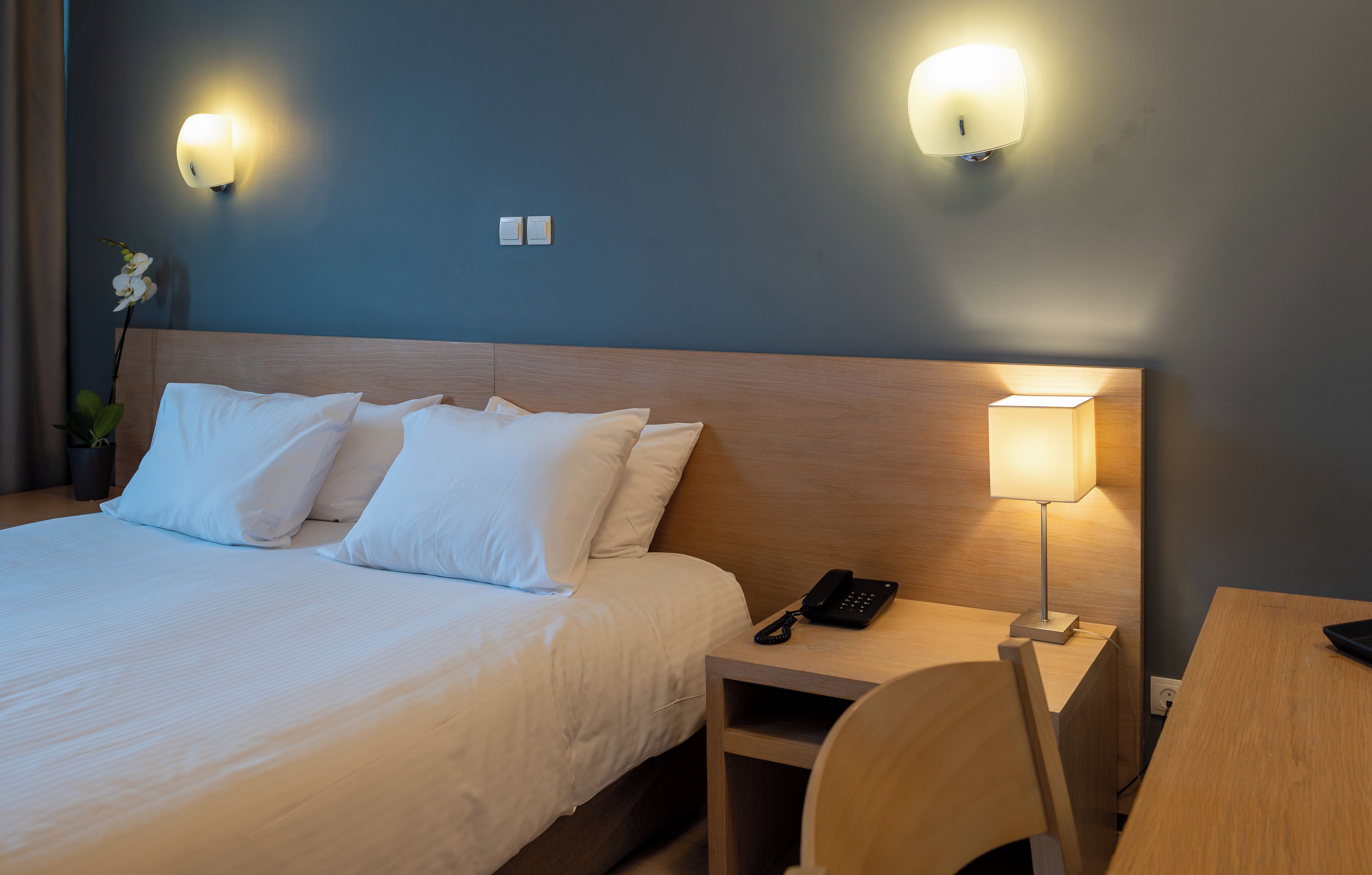 A hotel room with a bed, bedside table, lamp, phone, and a chair.