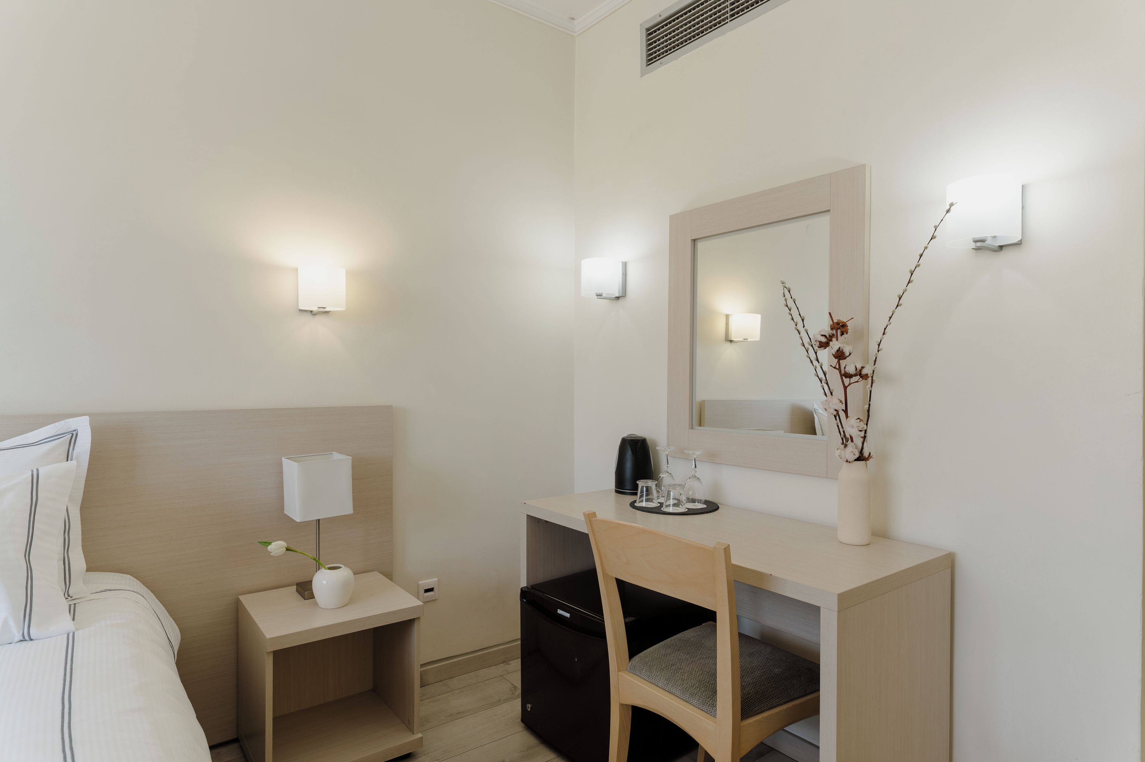 A hotel room with a bed, bedside table with a lamp, a desk with a mirror, a chair, and a small cabinet.
