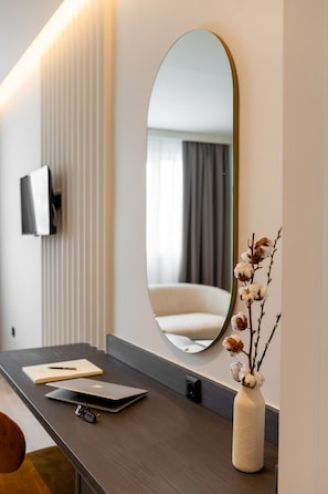 Une chambre d’hôtel moderne avec un bureau en bois foncé, un grand miroir ovale, un vase contenant des fleurs séchées et un téléviseur à écran plat fixé au mur.