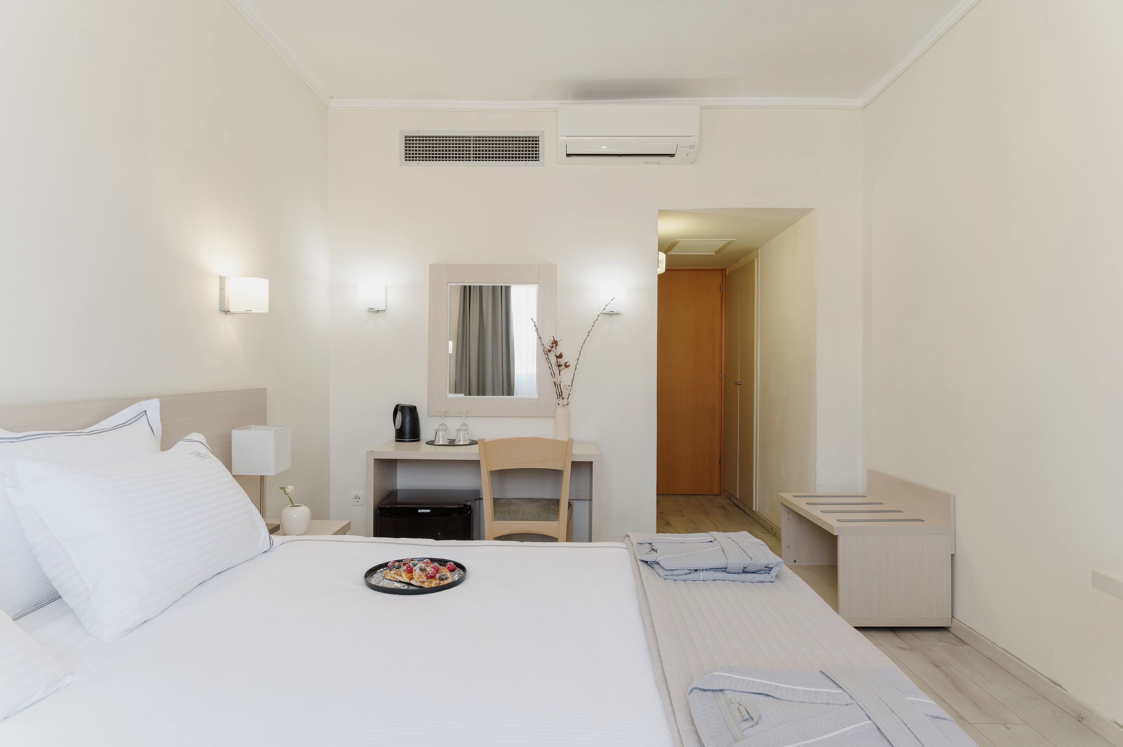 A hotel room with a bed, a desk, a chair, a small table with a lamp, a mirror, and a door leading to another room.