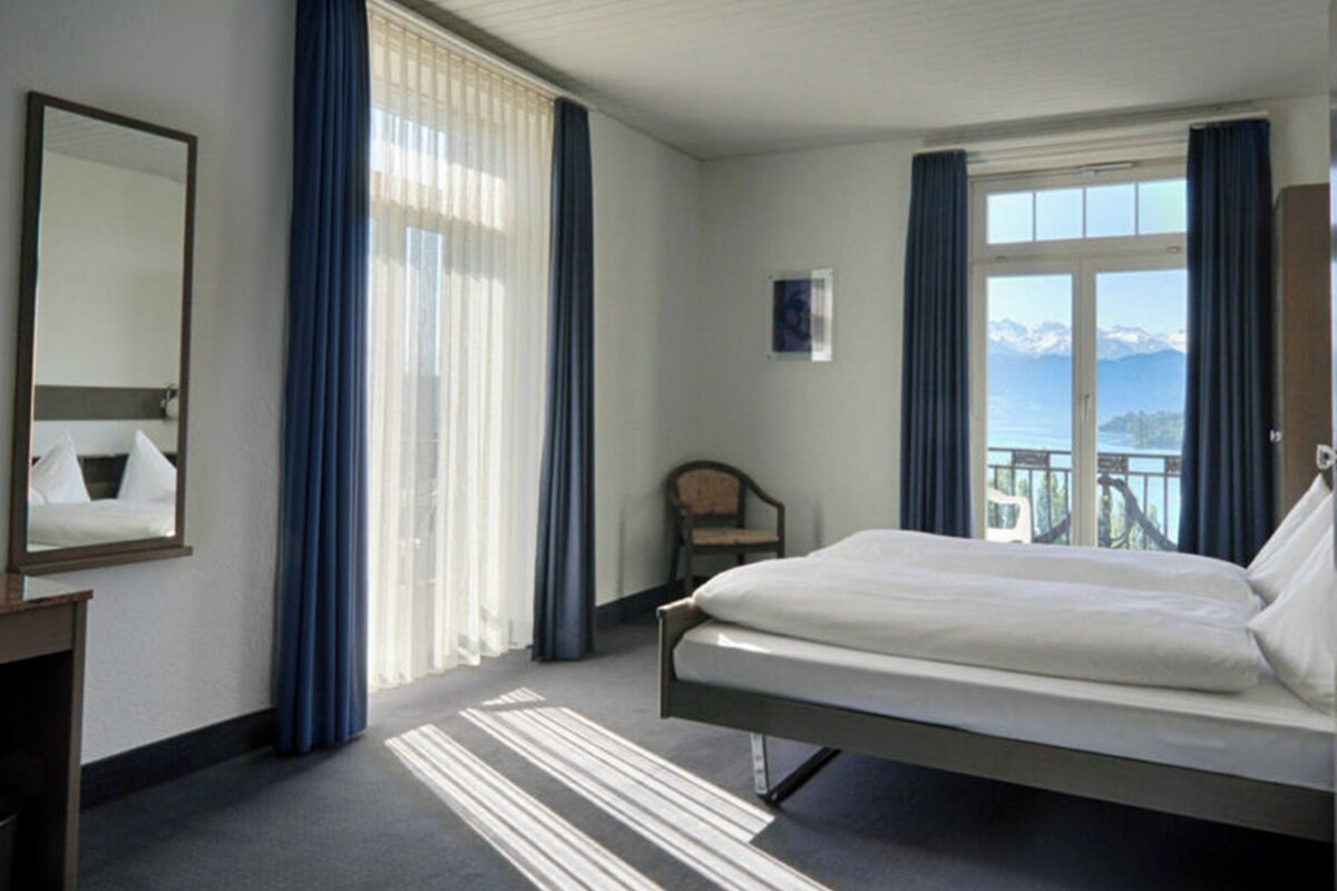 A hotel room with a bed, a chair, a desk, and a large window with a view of mountains.