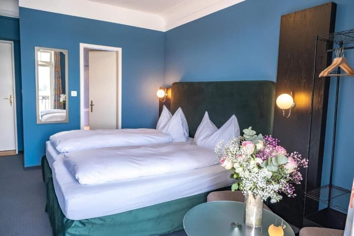 Une chambre d’hôtel avec un grand lit, une table de chevet ornée d’un vase de fleurs et une armoire contenant des vêtements suspendus.