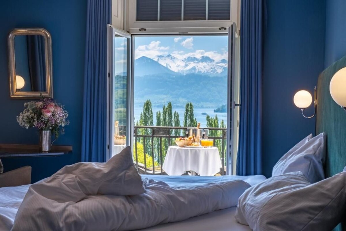 A hotel room with a bed, a bedside table with a vase of flowers, a balcony with a table set for breakfast, and a view of mountains and a lake.