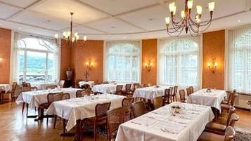 Une salle à manger spacieuse, dotée de nombreuses tables dressées, de grandes fenêtres laissant entrer la lumière naturelle et d’élégants lustres.