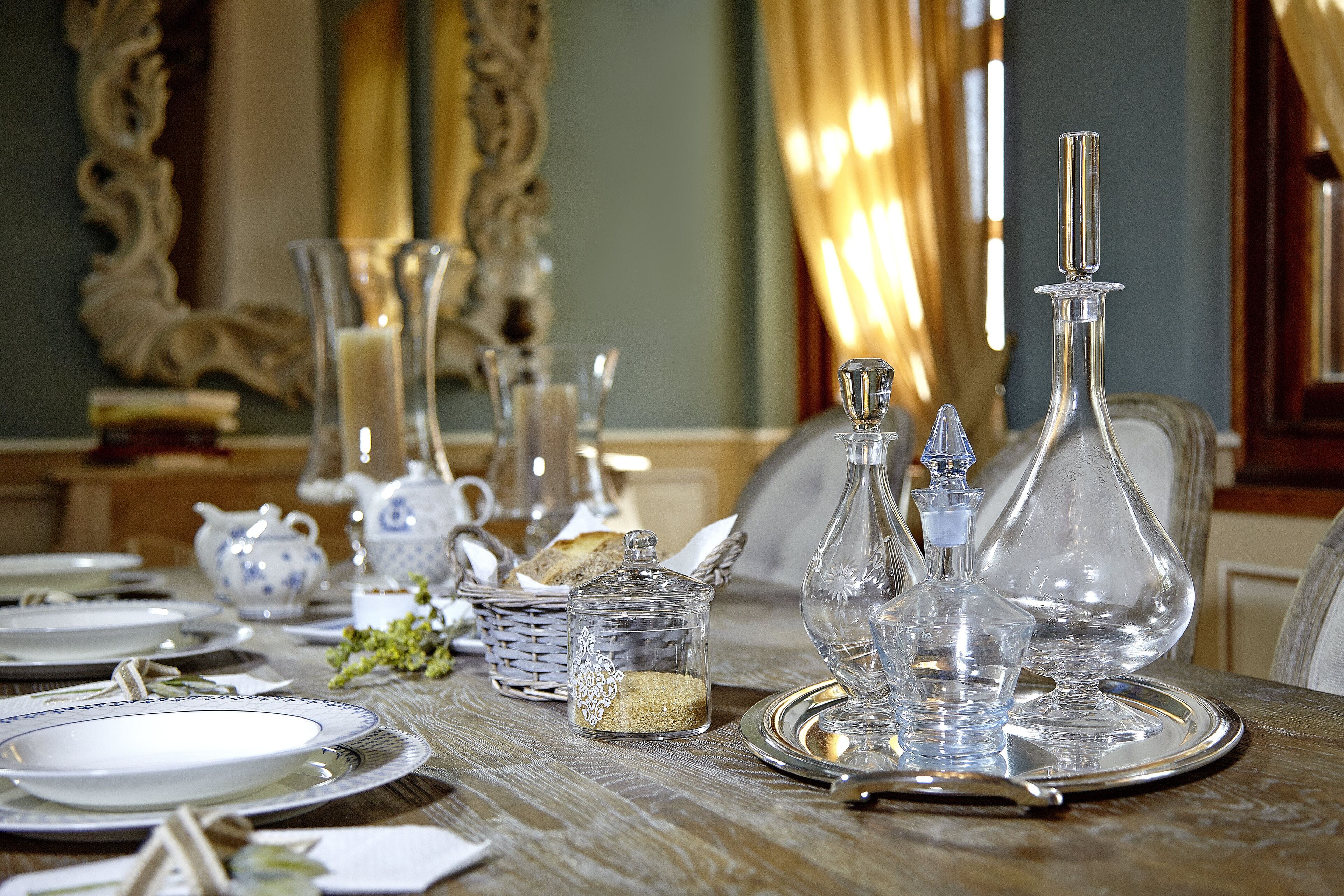 Une table à manger dressée avec de la vaisselle en verre transparent, des couverts en argent et des assiettes, agrémentée d’un miroir décoratif et de rideaux en arrière-plan.