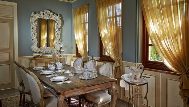 Comedor con una mesa de madera dispuesta para cuatro, un espejo grande y ornamentado, y cortinas doradas.