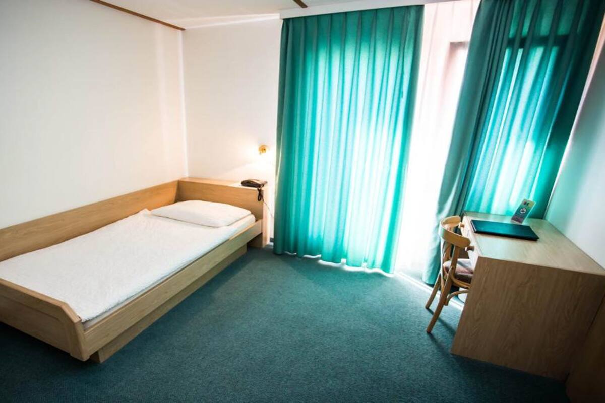 A small, single-bed room with a wooden headboard, a desk with a chair, a nightstand, and a window with teal curtains.