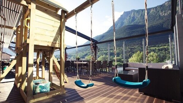 An outdoor deck with a wooden swing set, seating area, and a view of a mountainous landscape. - Hotel Olympica (Brig)
